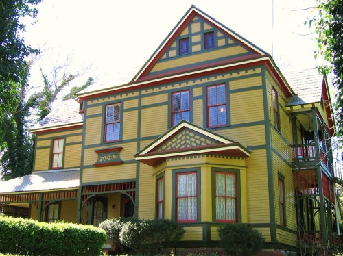 A colorful Victorian-style house with yellow siding, green trim, and red accents, surrounded by greenery.