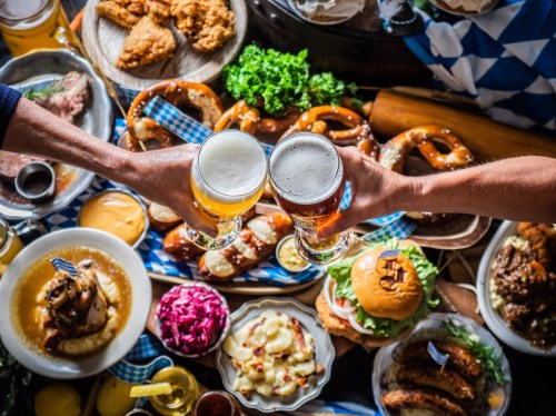 Two hands clink beer glasses over a festive spread of various foods, including pretzels, fried chicken, and sausages.