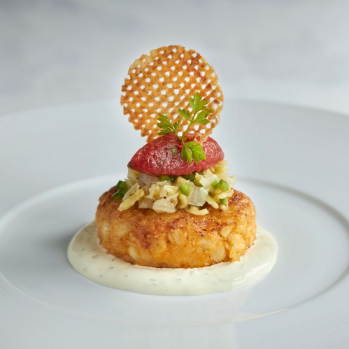 A gourmet dish featuring a crispy potato cake topped with crab salad, a slice of tomato, and a decorative wafer.