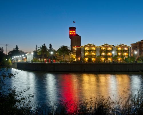 Napa Mill building illuminated at dusk, reflecting in the calm water of the river, surrounded by palm trees.