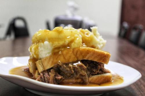 A plate with a sandwich made of bread, pulled meat, mashed potatoes, and gravy, served on a wooden table.
