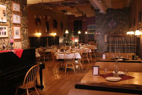 Cozy restaurant interior with wooden tables, chairs, and stone walls, featuring warm lighting and a piano in the corner.
