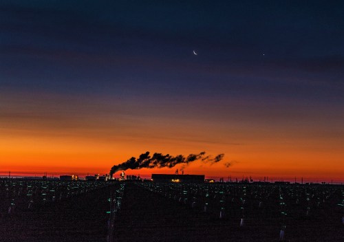 Silhouetted landscape at sunset with smoke rising from buildings and a crescent moon in the sky above.