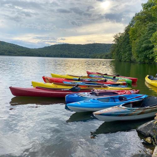 Colorful kayaks lined up on the shore of a calm lake, surrounded by lush green hills under a cloudy sky.