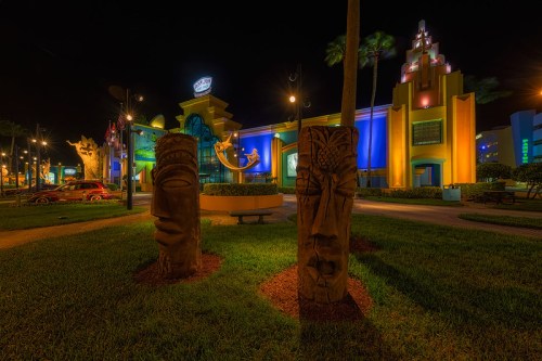 Colorful night scene of a building with tiki sculptures in the foreground, surrounded by palm trees and vibrant lights.