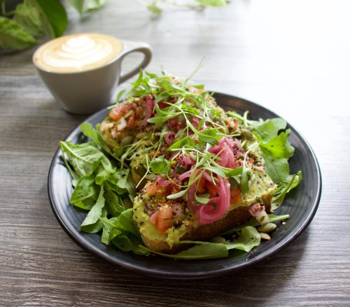 A plate of avocado toast topped with tomatoes, pickled onions, and microgreens, served with a latte on the side.