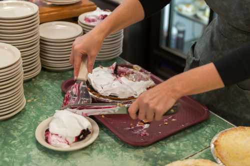 A person slices a pie and serves a portion onto a plate, with whipped cream topping and empty plates in the background.