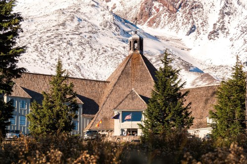 A mountain lodge with a distinctive peaked roof, surrounded by evergreen trees and snow-capped mountains in the background.