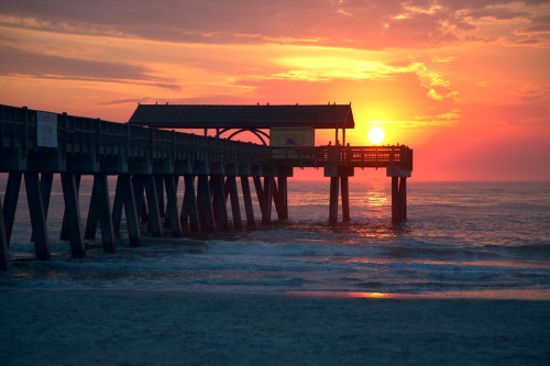 Silhouette of a pier at sunset, with vibrant orange and pink hues reflecting on the water.
