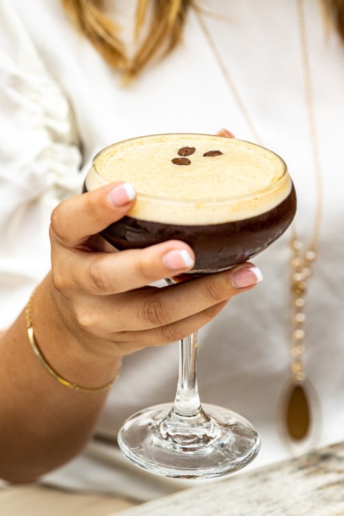A close-up of a hand holding a cocktail glass filled with a dark drink, topped with coffee beans.