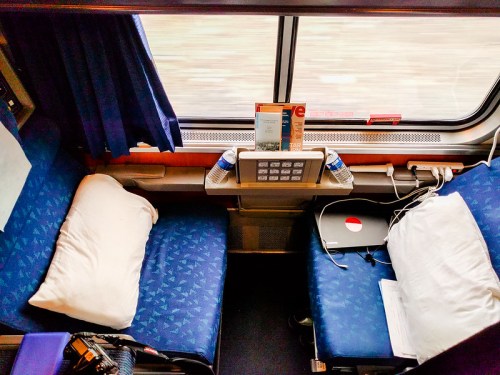 Interior of a train compartment with two seats, pillows, a table, and a laptop, viewed from above.