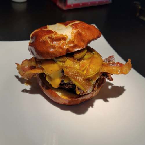 A pretzel bun burger topped with melted cheese, crispy bacon, and sautéed onions on a white plate.