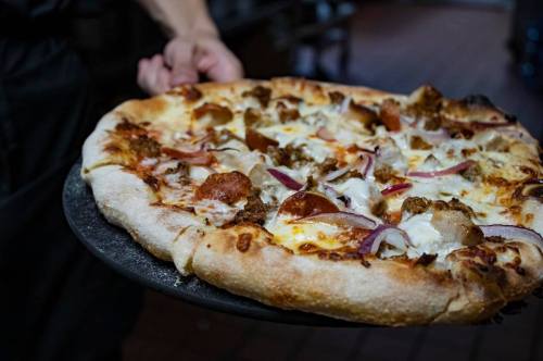 A person holds a freshly baked pizza topped with sausage, pepperoni, onions, and melted cheese on a black pizza tray.