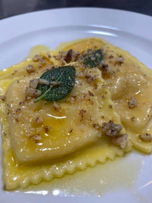 Two ravioli topped with sage leaves and crushed nuts, drizzled with a golden sauce, served on a white plate.