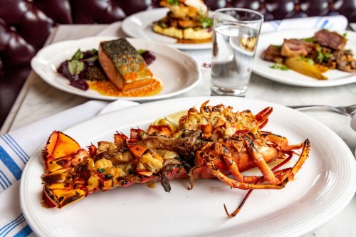 A plate of grilled lobster with lemon, accompanied by a glass of water and a blurred background of other dishes.