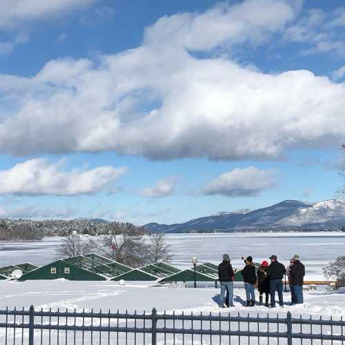 A group of people stands by a snowy lake, with mountains and clouds in the background under a clear blue sky.