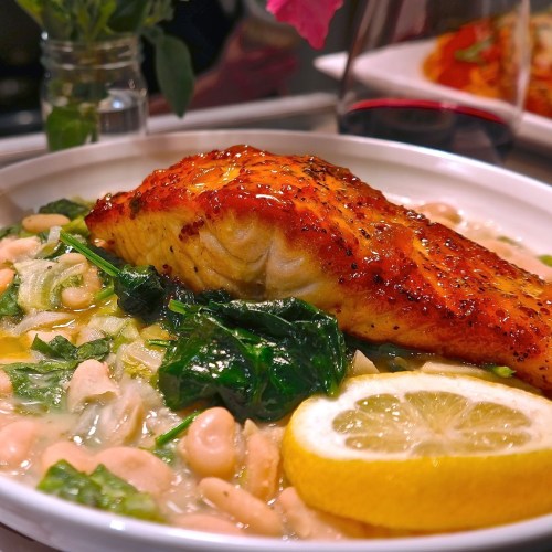 A plate of salmon on a bed of white beans and spinach, garnished with a lemon slice and herbs.