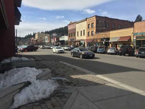 A bustling street scene with shops, parked cars, and snow along the sidewalk, set against a backdrop of mountains.