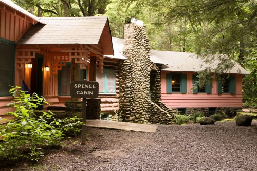 A rustic cabin with a stone chimney, surrounded by greenery, featuring a sign that reads "Spence Cabin."