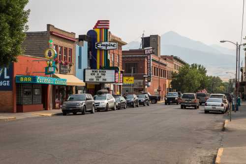 A street scene featuring vintage storefronts, a theater marquee, and parked cars, with mountains in the background.