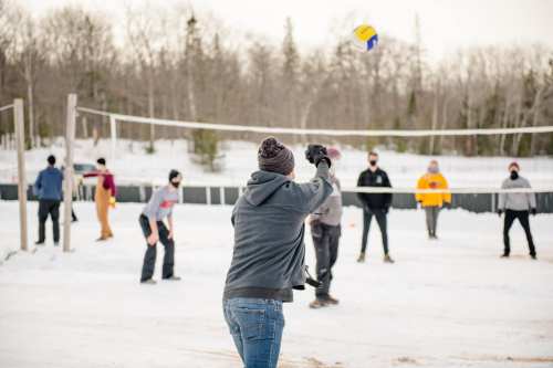A group of people playing volleyball on a snowy court, wearing winter clothing and masks.