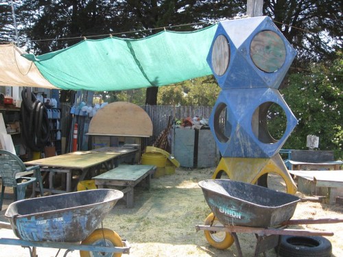 A colorful play structure surrounded by wheelbarrows and wooden tables under a green canopy in a yard.