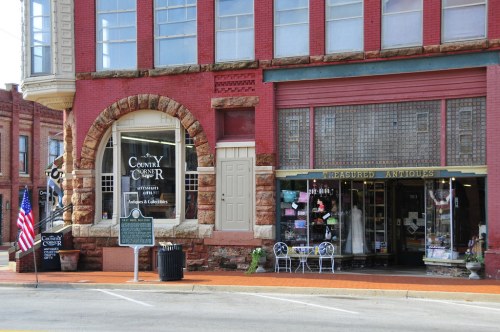 A red brick building featuring two shops: "Country Corner" and "Treasured Antiques," with a historical marker nearby.