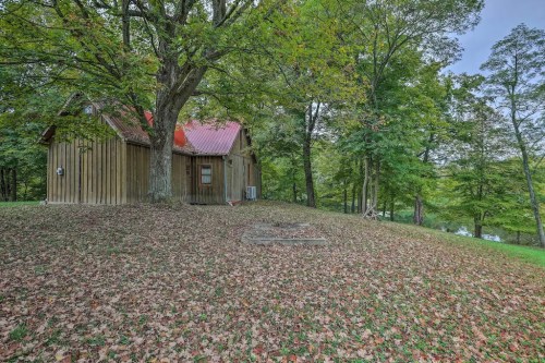 A rustic wooden cabin with a red roof, surrounded by trees and fallen leaves, near a calm body of water.