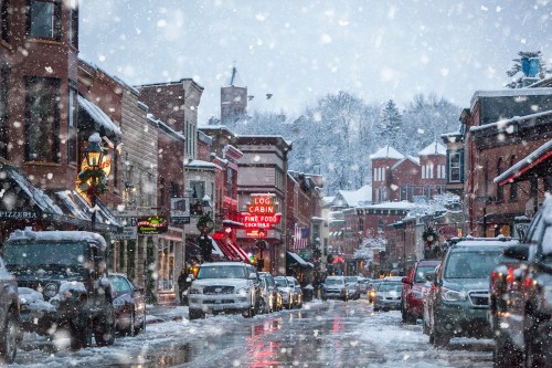 Snowy street scene with charming shops and restaurants, festive decorations, and cars parked along the road.