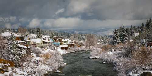 A snowy landscape with houses along a river, surrounded by trees and mountains under a cloudy sky.