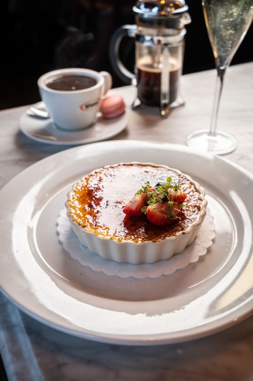 A creamy crème brûlée topped with strawberries, served on a white plate with coffee and a glass of sparkling wine in the background.