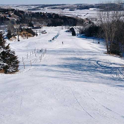 A snowy landscape with ski slopes, trees, and a distant figure on the hill, under a clear blue sky.