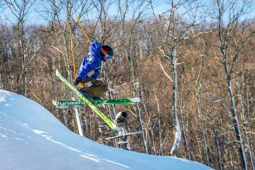 A skier in a blue jacket performs a jump over a snowy slope, surrounded by trees in winter.