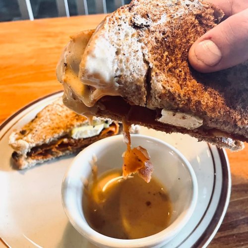 A hand holds a toasted sandwich with melted cheese, dripping, next to a small bowl of dipping sauce on a plate.