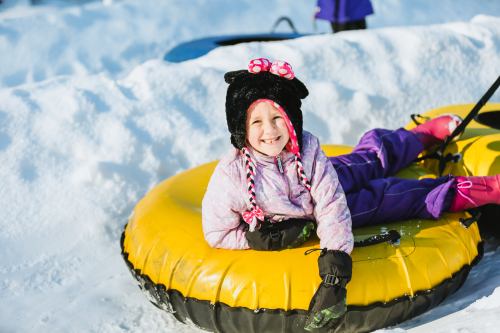 A smiling child in a pink and purple outfit relaxes on a yellow snow tube, wearing a black bear hat in a snowy setting.