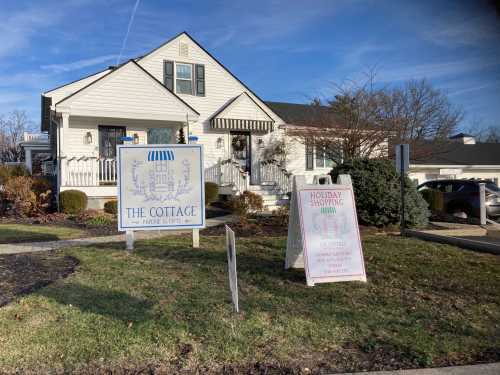 A white cottage with a sign reading "The Cottage" and a holiday shopping sign in front, surrounded by greenery.