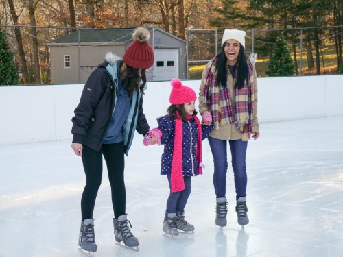 Three people ice skating together, two adults holding hands with a young girl in a pink hat and scarf, smiling.