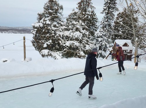 Two people skate on an outdoor ice rink surrounded by snow-covered trees and festive lights.