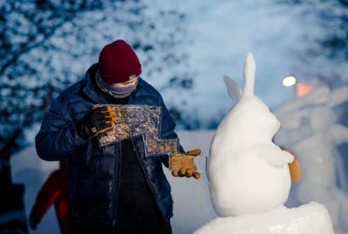 A person carves a snow sculpture of a rabbit, wearing gloves and a beanie, with trees in the background.