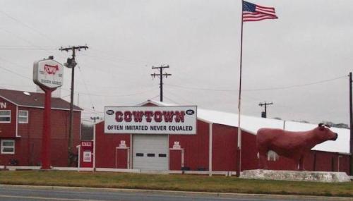A roadside view of Cowtown, featuring a large cow statue and a sign that reads "Often Imitated Never Equaled."