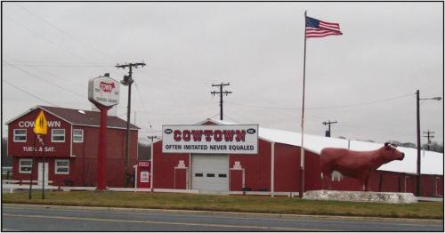 A roadside view of Cowtown, featuring a large cow statue, an American flag, and red buildings with signage.