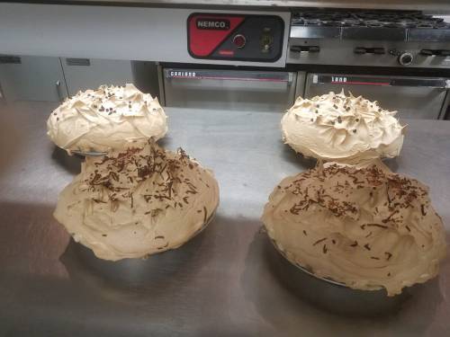 Four cream pies topped with chocolate shavings, placed on a stainless steel countertop in a kitchen.