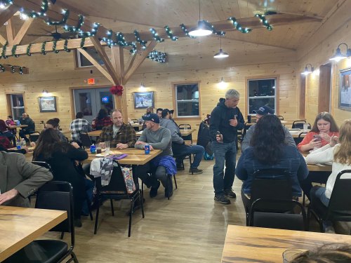 A cozy indoor gathering with people sitting at tables, chatting, and enjoying drinks in a warmly lit wooden space.