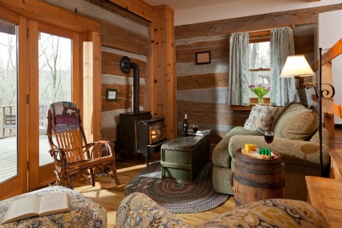 Cozy cabin interior with wooden walls, a rocking chair, a green sofa, a wood stove, and a view of nature outside.
