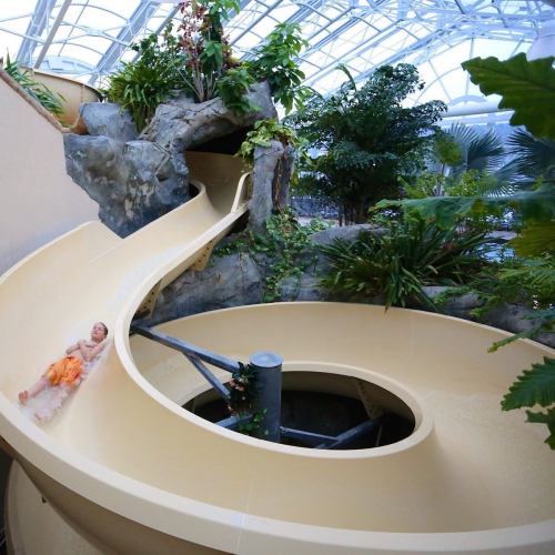 A water slide spirals down through a lush indoor garden, with a person enjoying the ride.