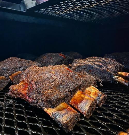 Close-up of smoked beef ribs on a grill, showcasing a dark, flavorful crust and tender meat.