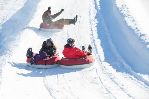 Three people enjoy snow tubing down a snowy slope, laughing and having fun on red inflatable tubes.