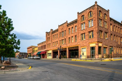 Historic buildings line a quiet street, showcasing red stone architecture and storefronts under a cloudy sky.