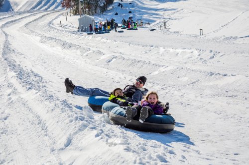 A man and two children enjoy tubing down a snowy hill on a sunny day, with others playing in the background.