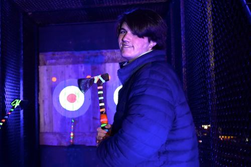 A person in a blue jacket holds a colorful axe in an illuminated axe-throwing venue, smiling at the camera.